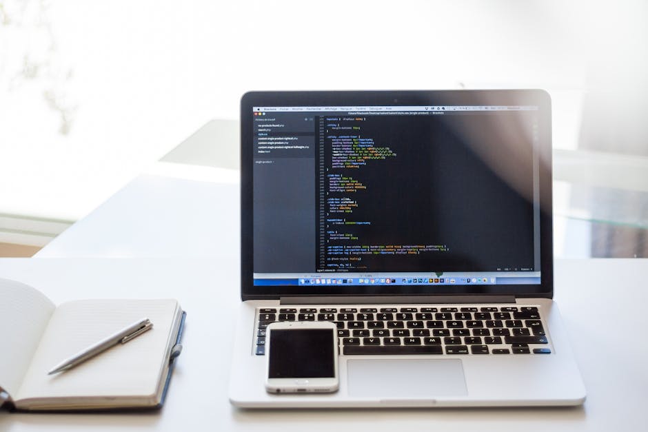 A silver laptop open on a white desk displaying lines of computer code in a dark-themed development environment, with a white smartphone resting on the laptop's keyboard. To the left of the laptop, th